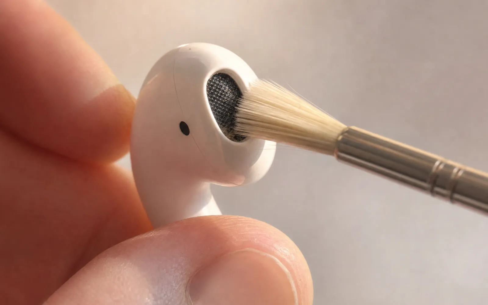 A wireless earbud held between fingertips with a soft dry brush against its speaker mesh